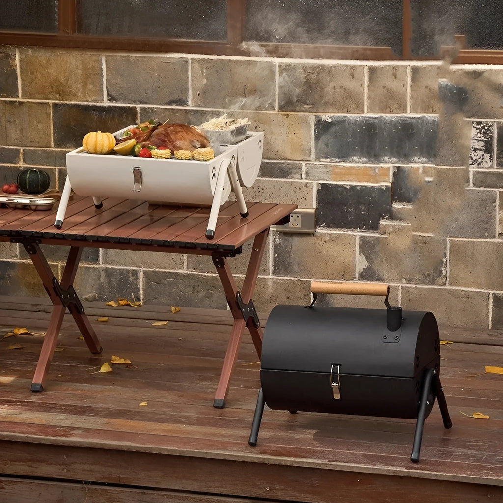 White portable charcoal barbecue grill with chimney oven, folded legs, two grill meshes, and detachable fuel tanks on a wooden table.