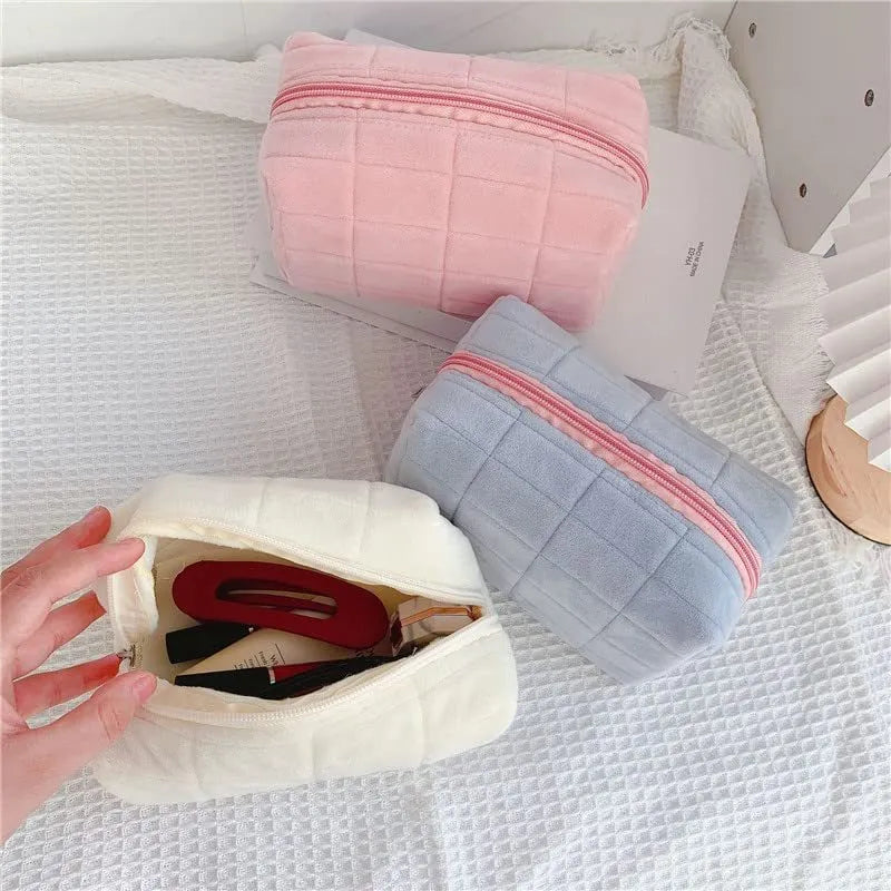 Three plush makeup bags in pink, white, and blue with zippers, laid flat showing front