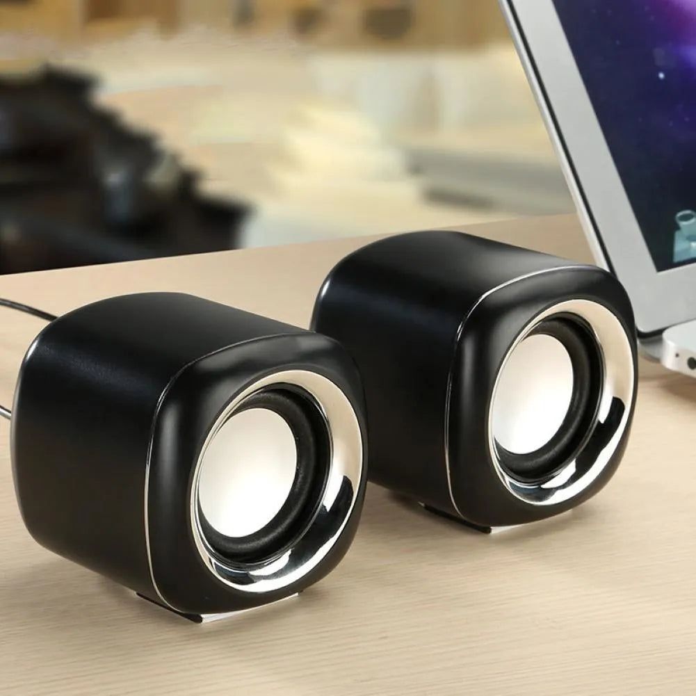 A pair of small black wired computer speakers with a volume scroll wheel, shown next to a 3.5mm audio plug on a desk.