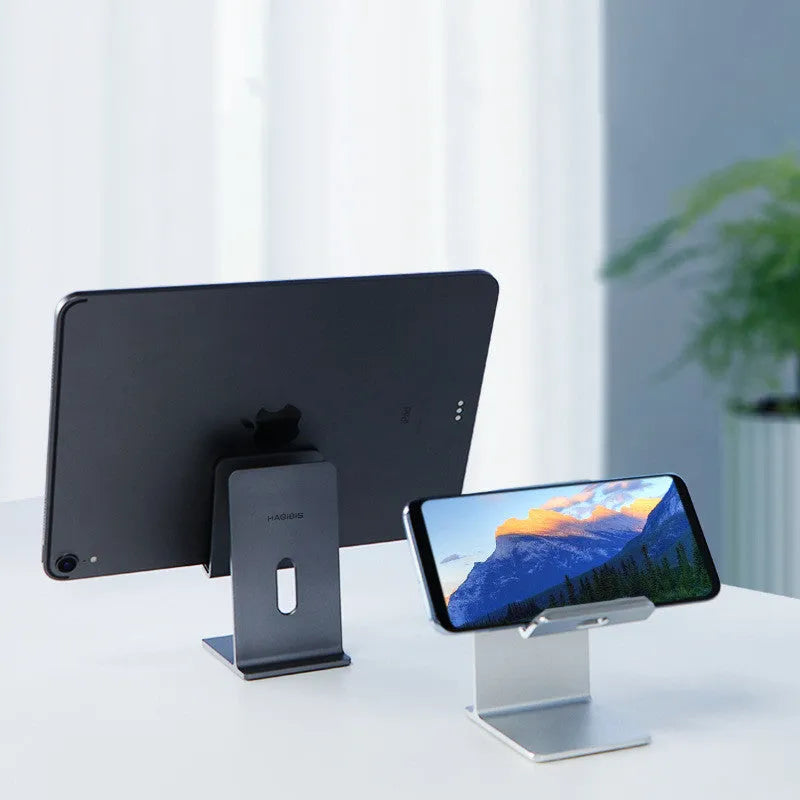 Dark space grey aluminium alloy mobile phone holder with adjustable clamp on a desk edge, holding a large tablet.