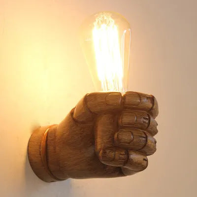 Close-up of a black resin fist sculpture wall lamp mounted on a dark wall, casting ambient light downwards.