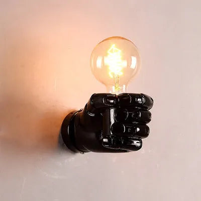 Close-up of a black resin fist sculpture wall lamp mounted on a dark wall, casting ambient light downwards.