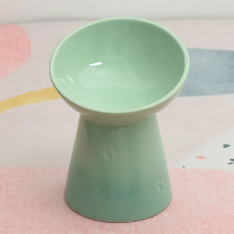 A high-footed white ceramic pet bowl with a slanted rim and non-slip black base, shown on a wooden floor.