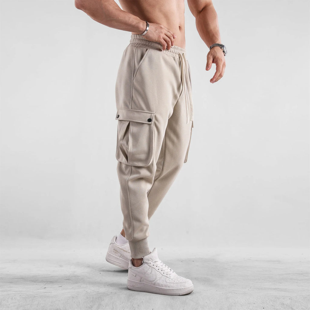 Men's loose-fit beige cotton trousers with tied ankle cuffs and multiple pockets on a neutral background.