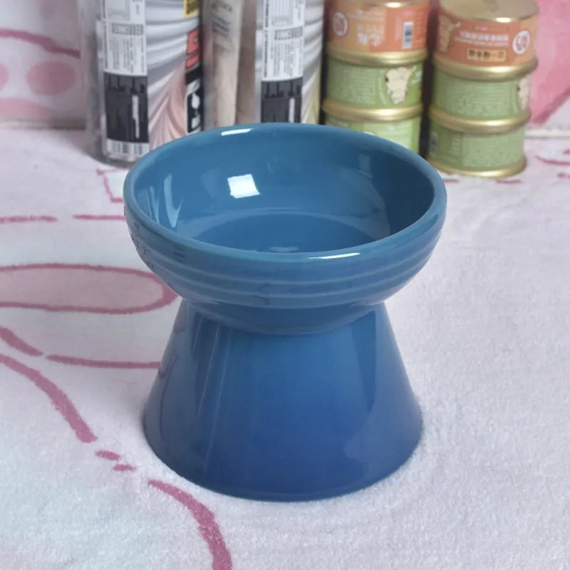 A high-footed white ceramic pet bowl with a slanted rim and non-slip black base, shown on a wooden floor.