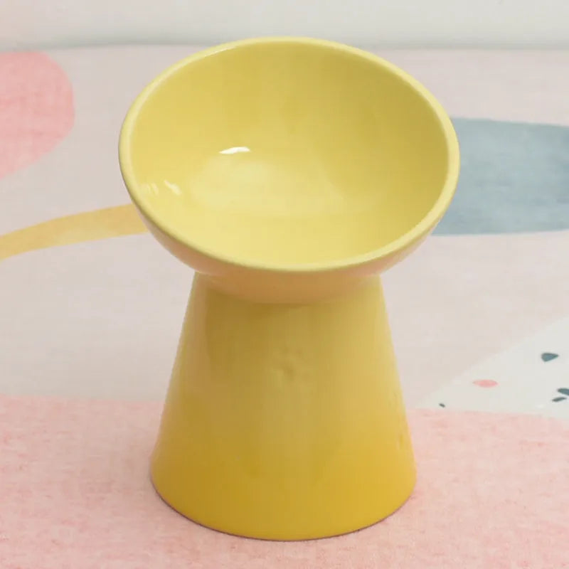 A high-footed white ceramic pet bowl with a slanted rim and non-slip black base, shown on a wooden floor.