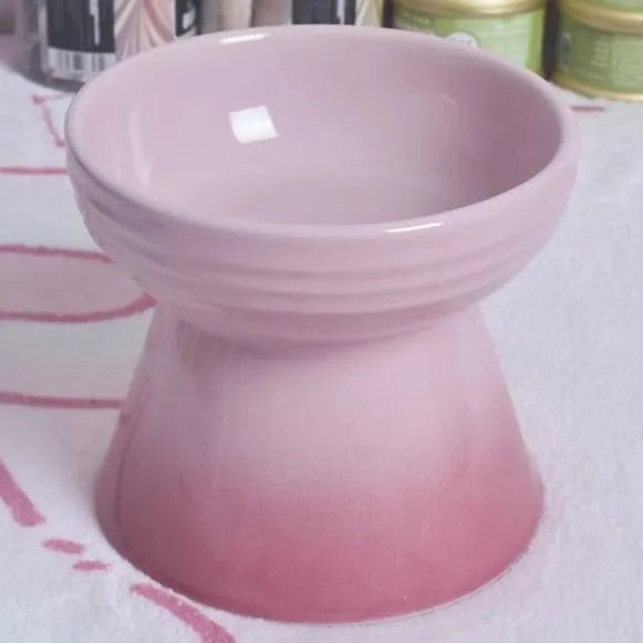 A high-footed white ceramic pet bowl with a slanted rim and non-slip black base, shown on a wooden floor.