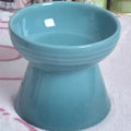 A high-footed white ceramic pet bowl with a slanted rim and non-slip black base, shown on a wooden floor.