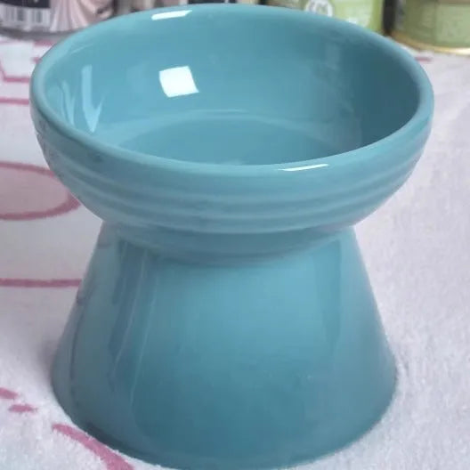A high-footed white ceramic pet bowl with a slanted rim and non-slip black base, shown on a wooden floor.