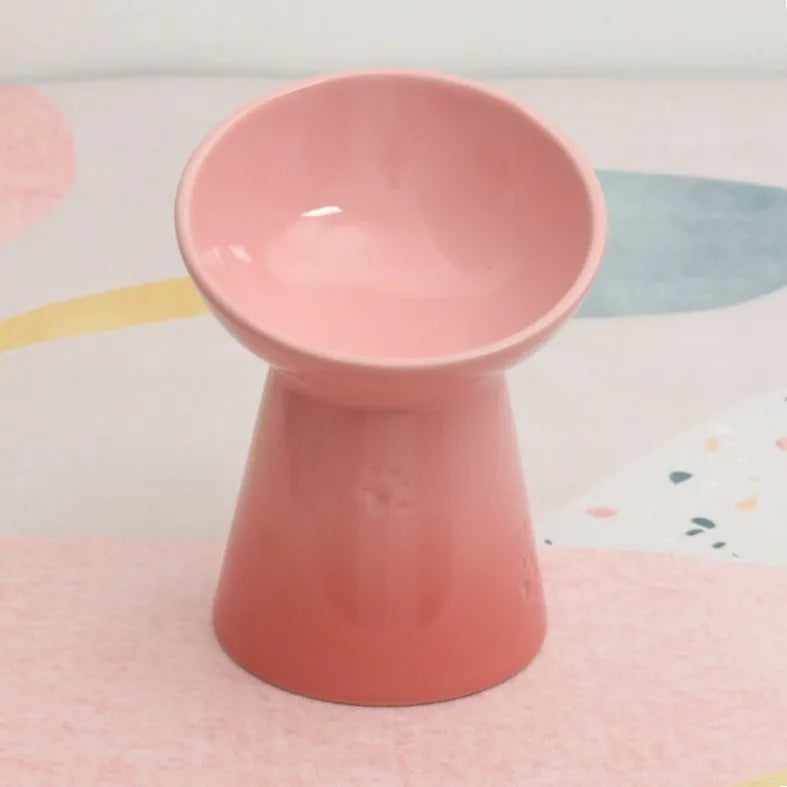 A high-footed white ceramic pet bowl with a slanted rim and non-slip black base, shown on a wooden floor.