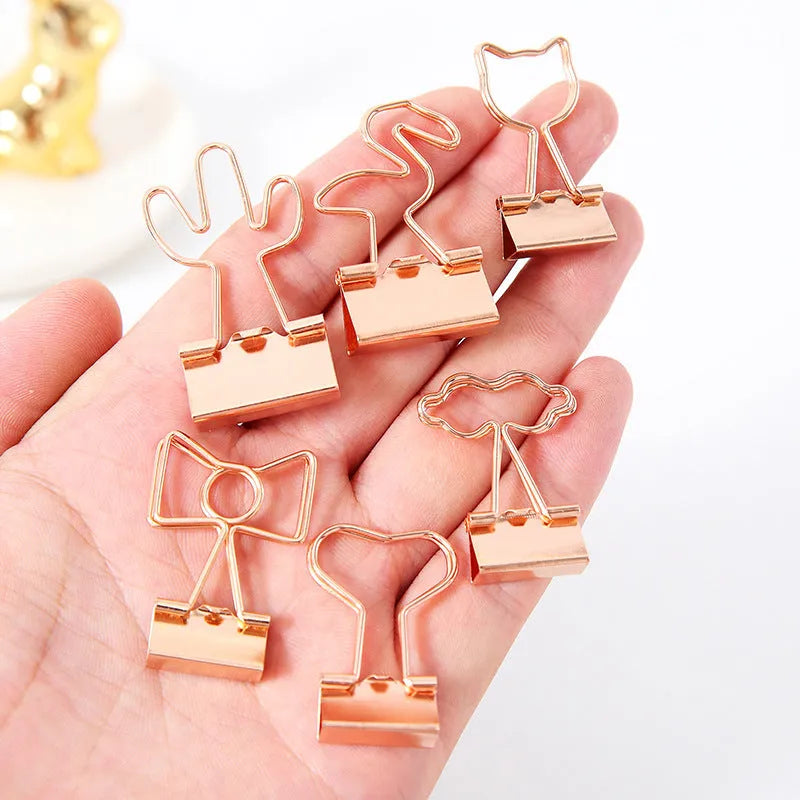 Small gold metal office clip with a long tail, shown in flamingo, love, cloud, cat, cactus, and bow shapes.