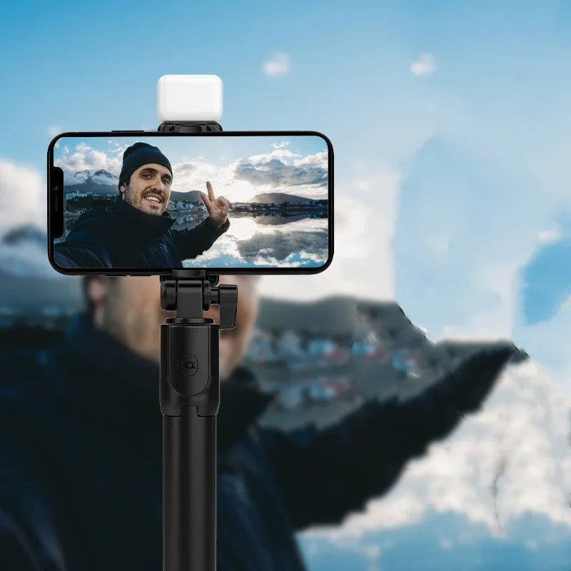 Black Bluetooth selfie stick with extended tripod legs and integrated fill light, holding a smartphone.