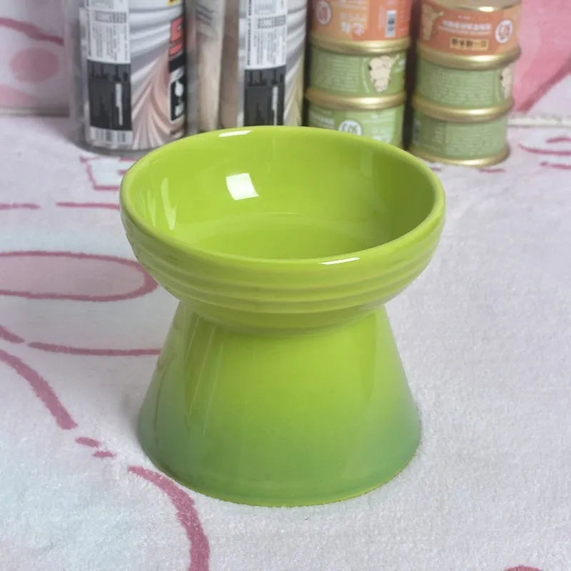 A high-footed white ceramic pet bowl with a slanted rim and non-slip black base, shown on a wooden floor.