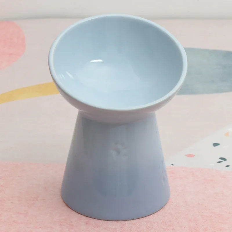 A high-footed white ceramic pet bowl with a slanted rim and non-slip black base, shown on a wooden floor.