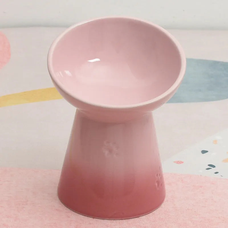 A high-footed white ceramic pet bowl with a slanted rim and non-slip black base, shown on a wooden floor.