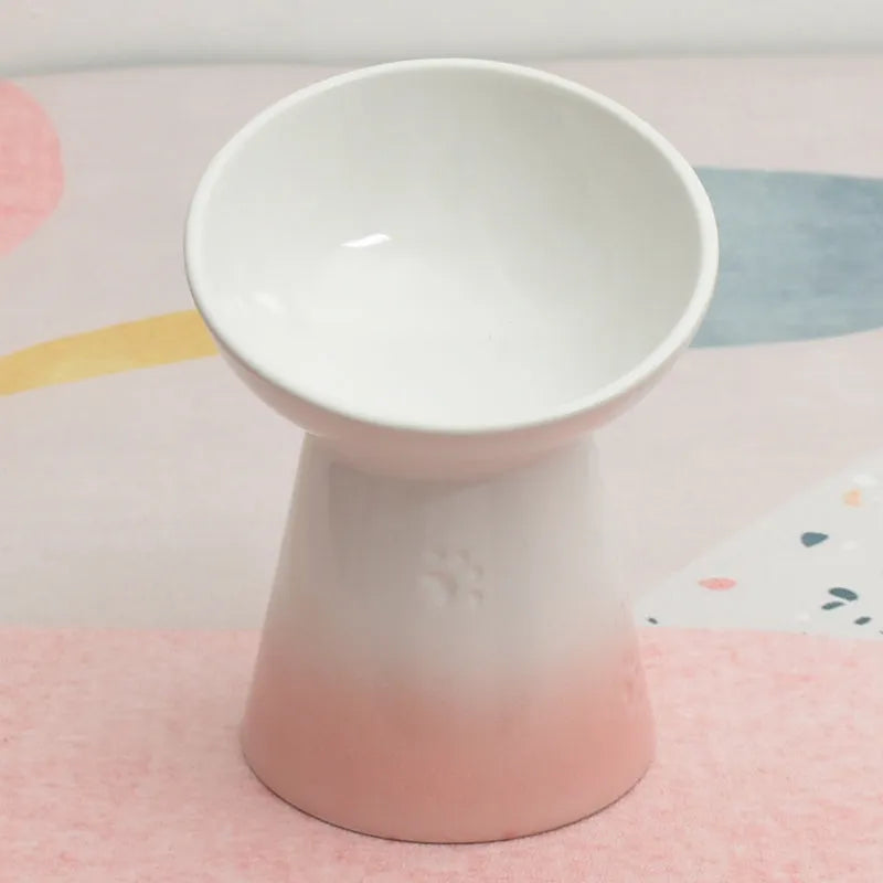 A high-footed white ceramic pet bowl with a slanted rim and non-slip black base, shown on a wooden floor.