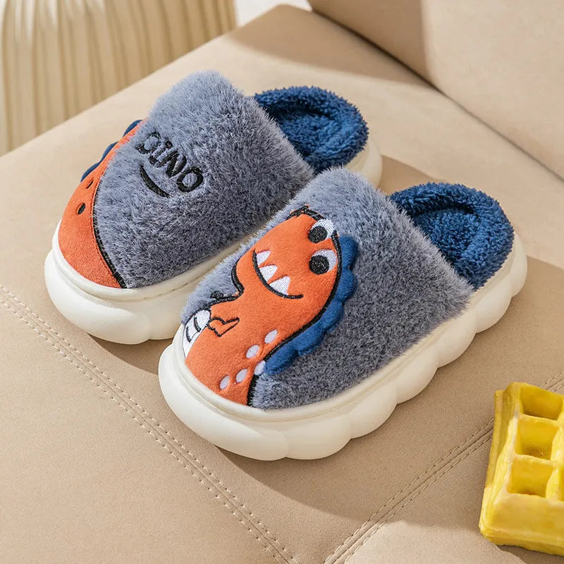 A pair of grey dinosaur-print plush slippers on a wooden floor, showing the soft cotton upper and thick textured sole.
