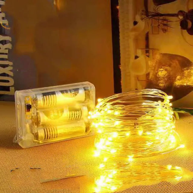 Close-up of bendable copper wire fairy lights with small LEDs, showing battery box and USB cable on a wooden surface.