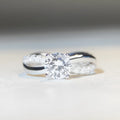 A silver ring with a 1 karat moissanite stone in a four-claw setting on a twisted geometric band.