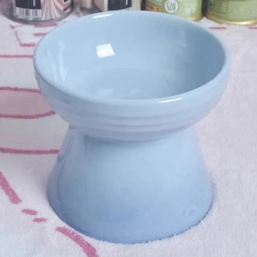 A high-footed white ceramic pet bowl with a slanted rim and non-slip black base, shown on a wooden floor.