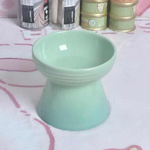 A high-footed white ceramic pet bowl with a slanted rim and non-slip black base, shown on a wooden floor.