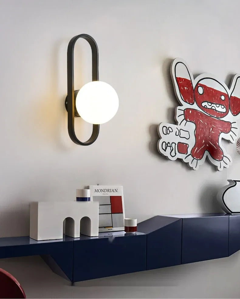 Minimalist circular wall lamp with glass shade on iron arm, in black finish, mounted beside a bed.
