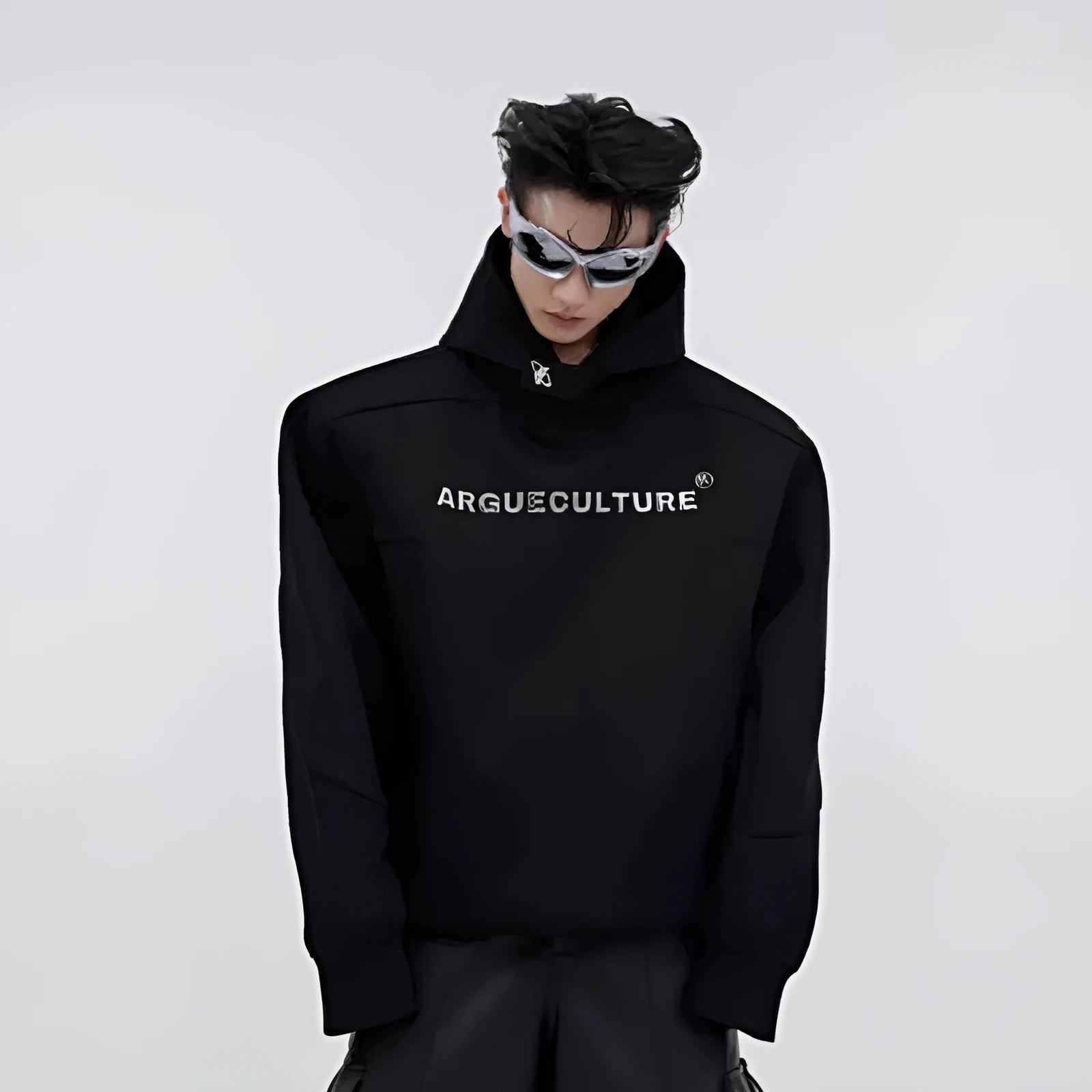 Man modelling a loose grey hooded sweater with heavy shoulder pads and raised metal-effect graphic printing on the front.