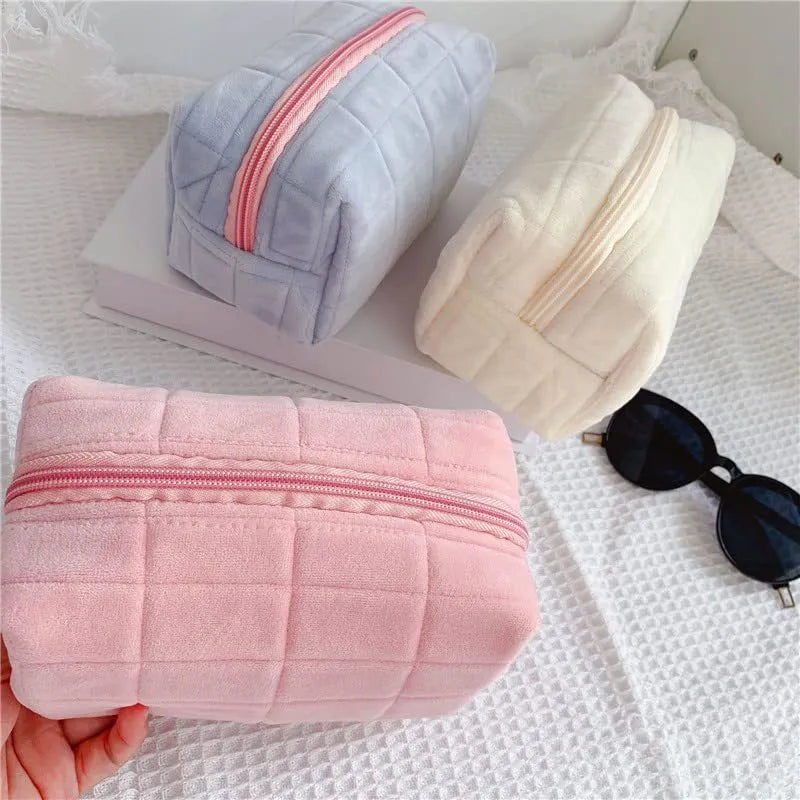 Three plush makeup bags in pink, white, and blue with zippers, laid flat showing front