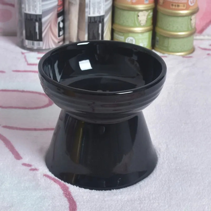 A high-footed white ceramic pet bowl with a slanted rim and non-slip black base, shown on a wooden floor.
