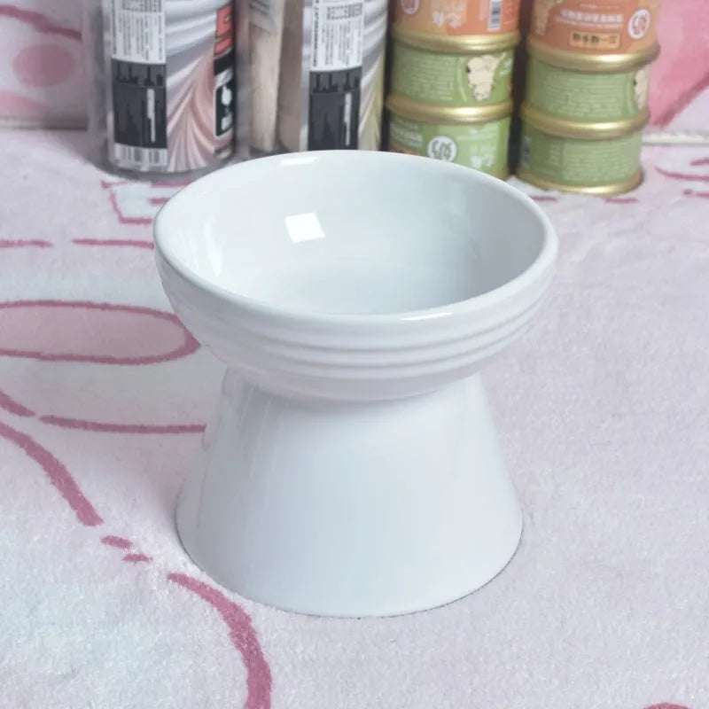 A high-footed white ceramic pet bowl with a slanted rim and non-slip black base, shown on a wooden floor.
