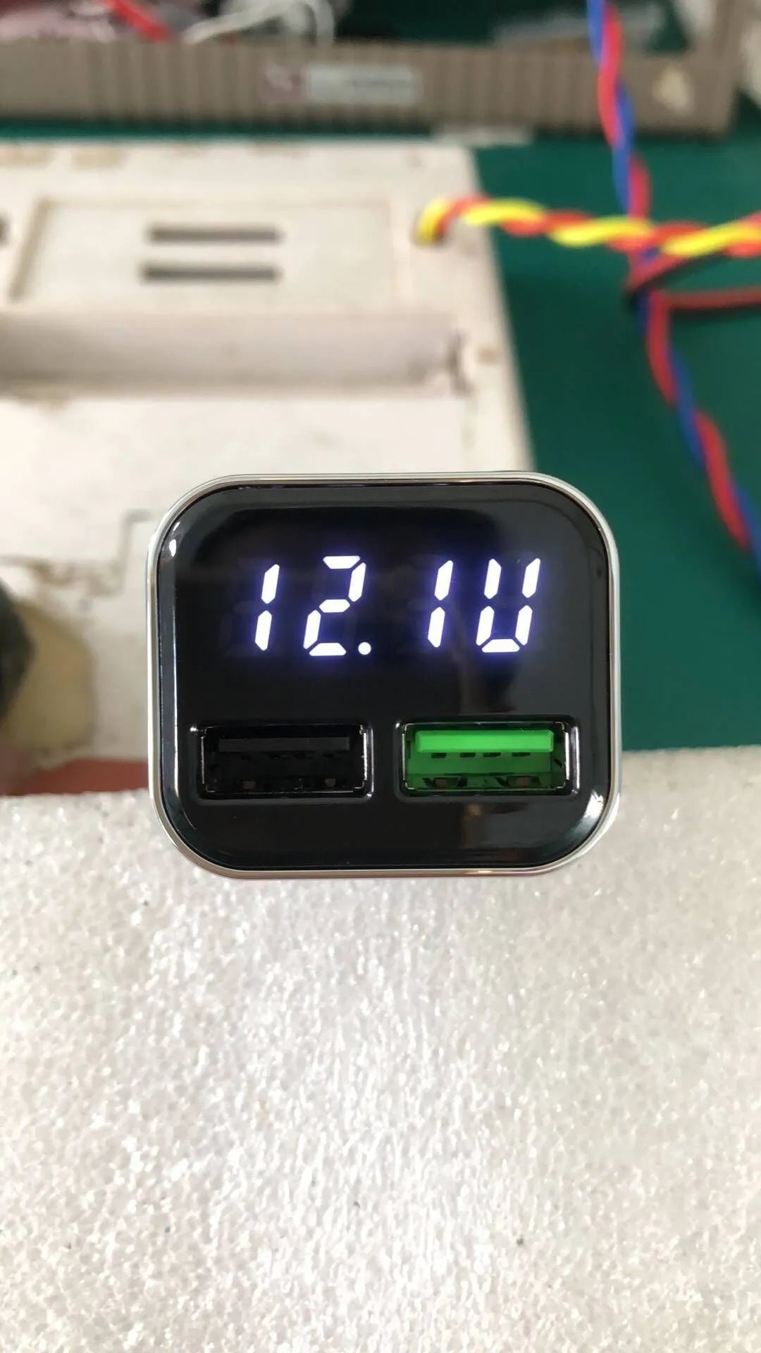 Black dual USB car charger with LCD screen displaying voltage, plugged into a car dashboard socket.