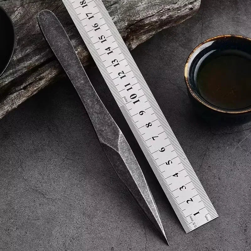 Black stainless steel hand pry tea knife on dark surface with tea leaves