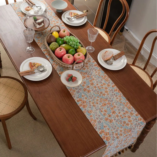 Beige jacquard table runner with floral leaf pattern and long tassels draped on a wooden dining table.