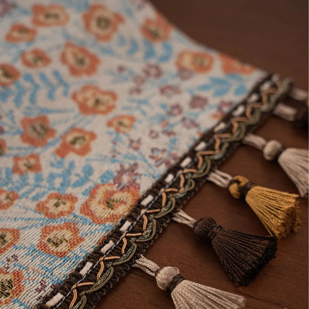 Beige jacquard table runner with floral leaf pattern and long tassels draped on a wooden dining table.