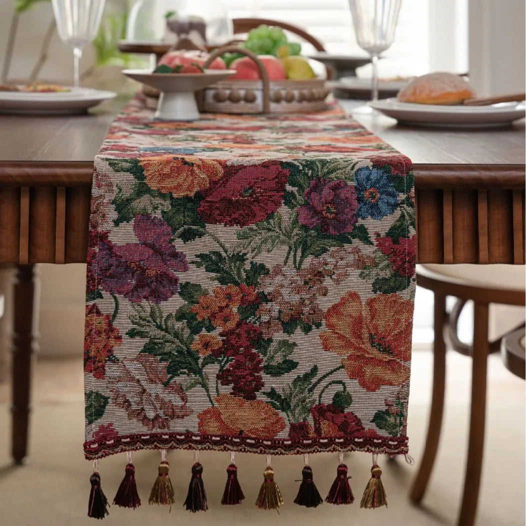 Beige jacquard table runner with floral leaf pattern and long tassels draped on a wooden dining table.