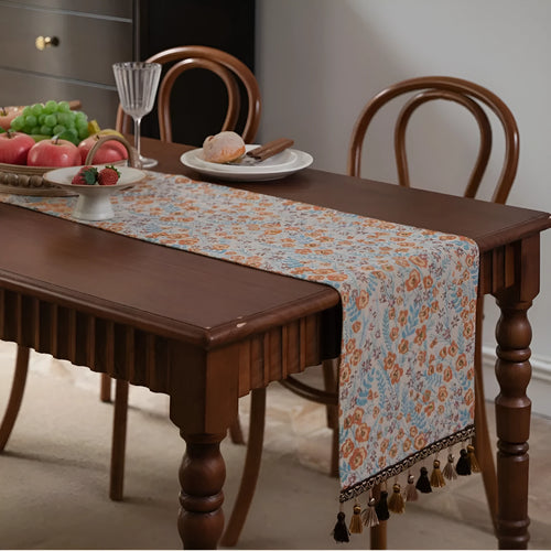 Beige jacquard table runner with floral leaf pattern and long tassels draped on a wooden dining table.