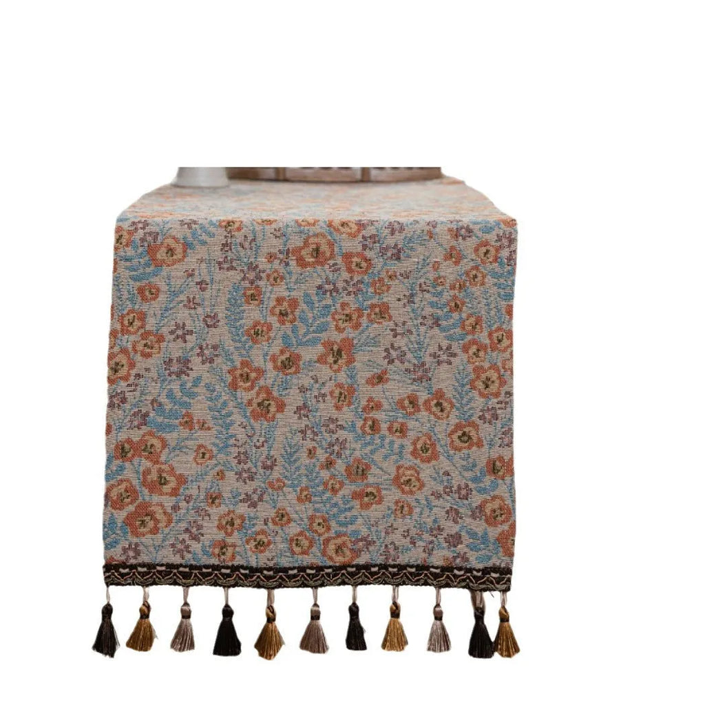 Beige jacquard table runner with floral leaf pattern and long tassels draped on a wooden dining table.
