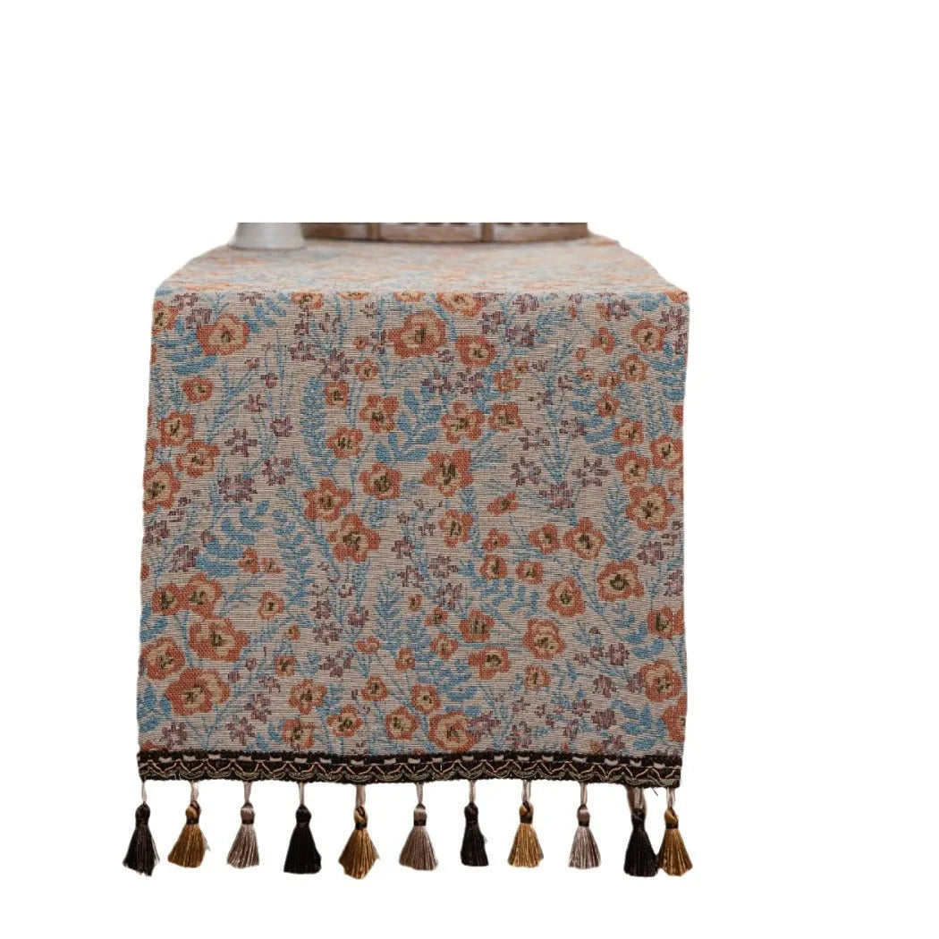Beige jacquard table runner with floral leaf pattern and long tassels draped on a wooden dining table.