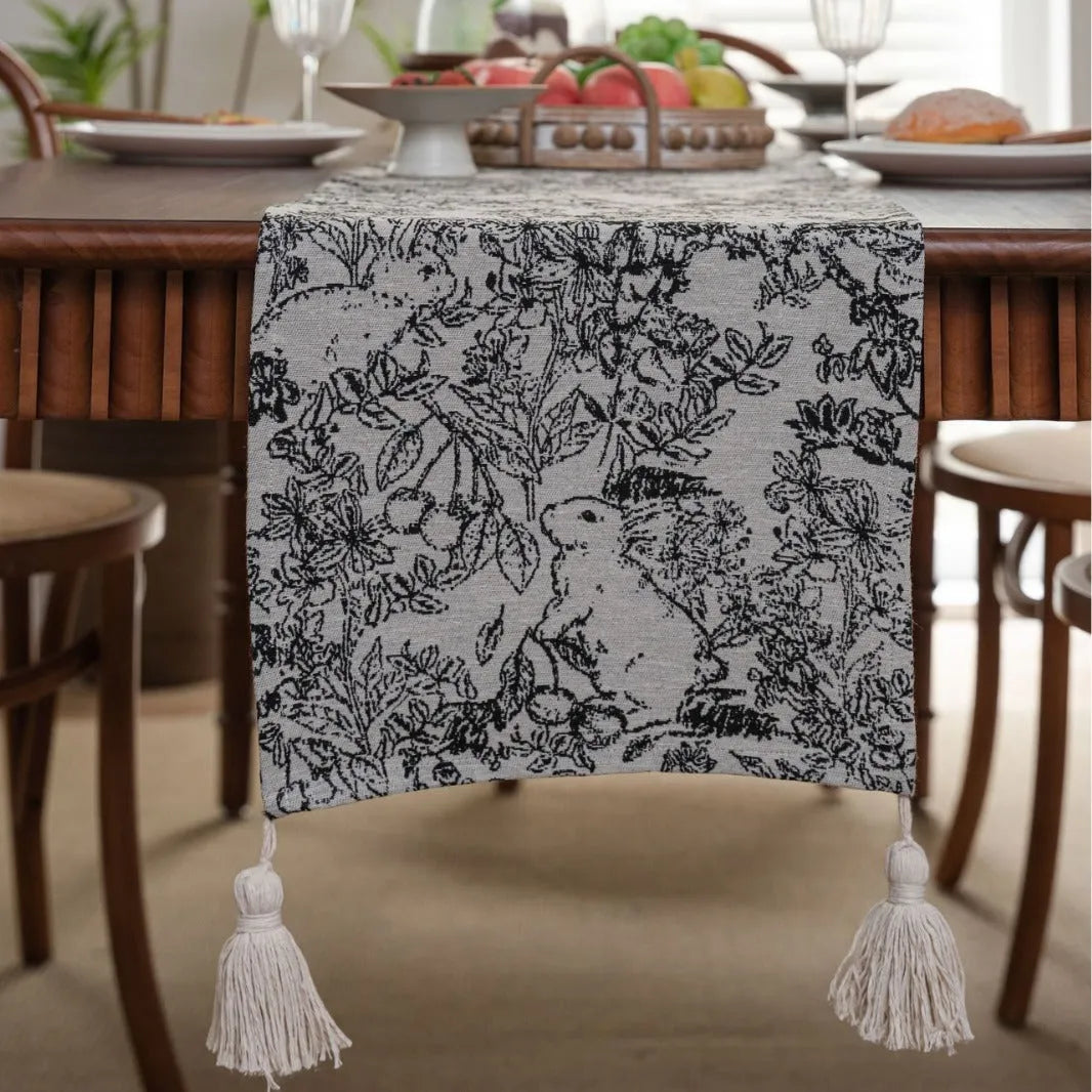 Beige jacquard table runner with floral leaf pattern and long tassels draped on a wooden dining table.