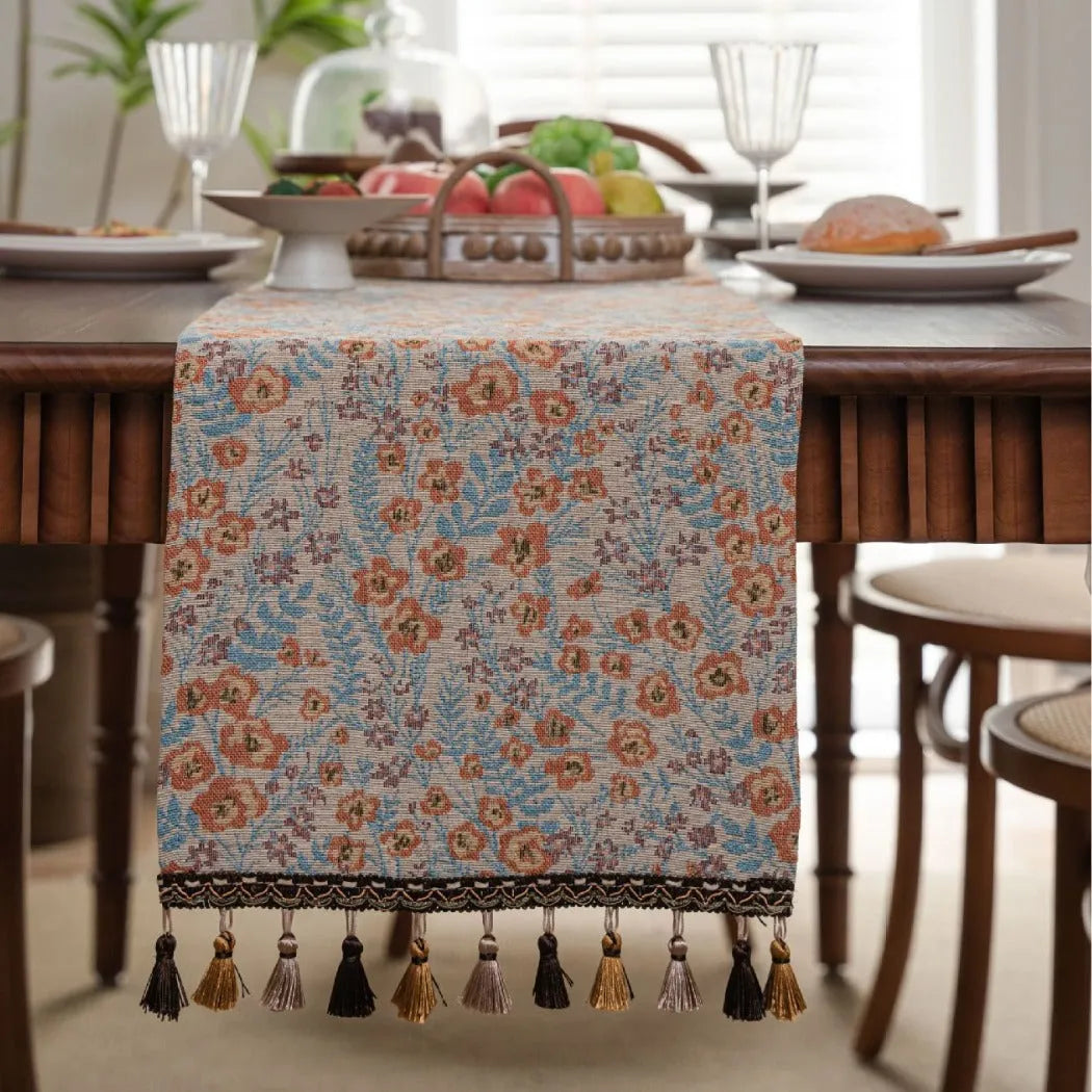 Beige jacquard table runner with floral leaf pattern and long tassels draped on a wooden dining table.