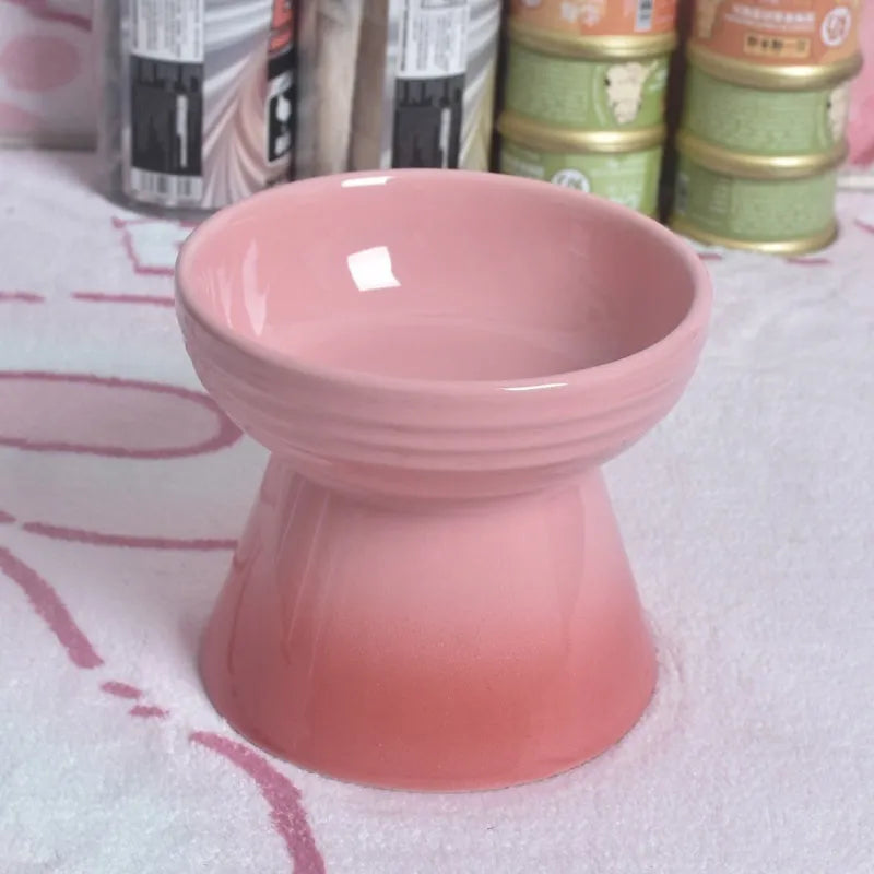 A high-footed white ceramic pet bowl with a slanted rim and non-slip black base, shown on a wooden floor.