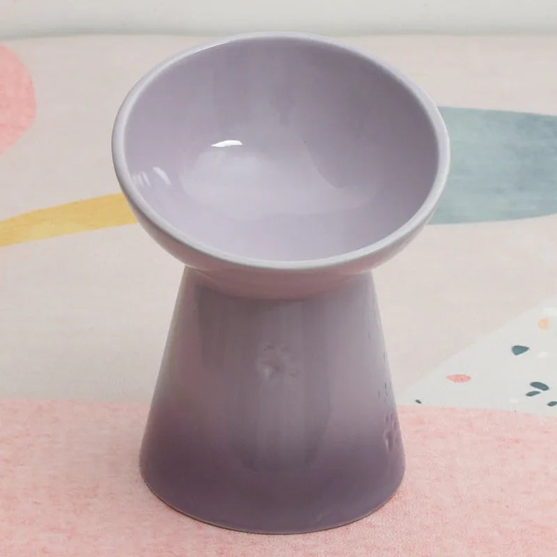 A high-footed white ceramic pet bowl with a slanted rim and non-slip black base, shown on a wooden floor.