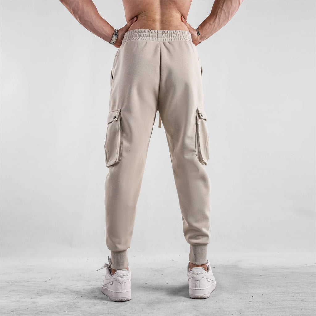 Men's loose-fit beige cotton trousers with tied ankle cuffs and multiple pockets on a neutral background.