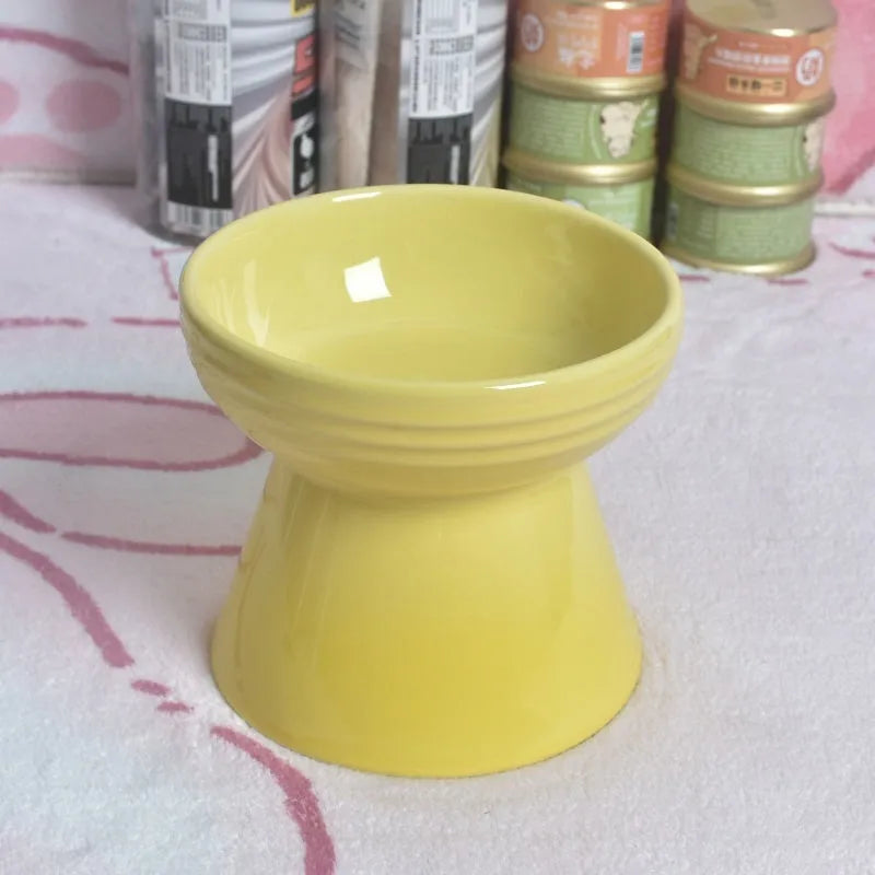 A high-footed white ceramic pet bowl with a slanted rim and non-slip black base, shown on a wooden floor.