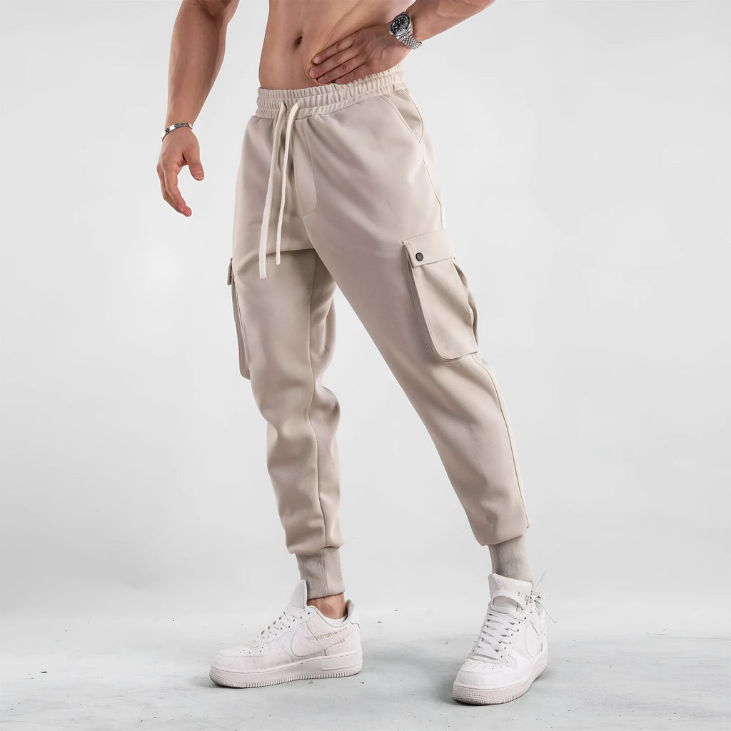 Men's loose-fit beige cotton trousers with tied ankle cuffs and multiple pockets on a neutral background.