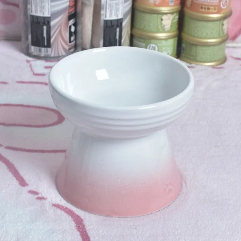 A high-footed white ceramic pet bowl with a slanted rim and non-slip black base, shown on a wooden floor.