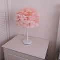 Handmade feather table lamp with metal base, white feathers, and remote control on a wooden bedside table