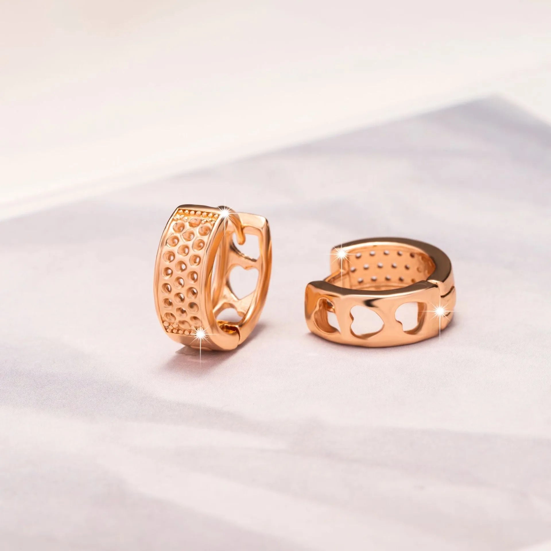 A pair of circular rose gold tone earrings featuring a hollowed-out heart shape design on a white background.