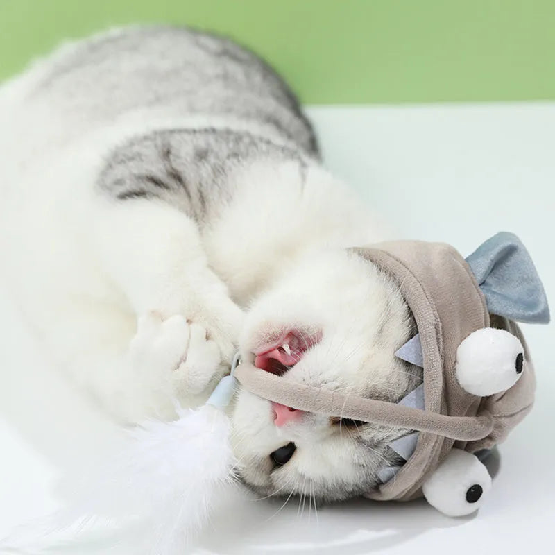 Grey dinosaur-themed cat headband toy with big eyes, a feather teaser, and an adjustable strap for hands-free play.