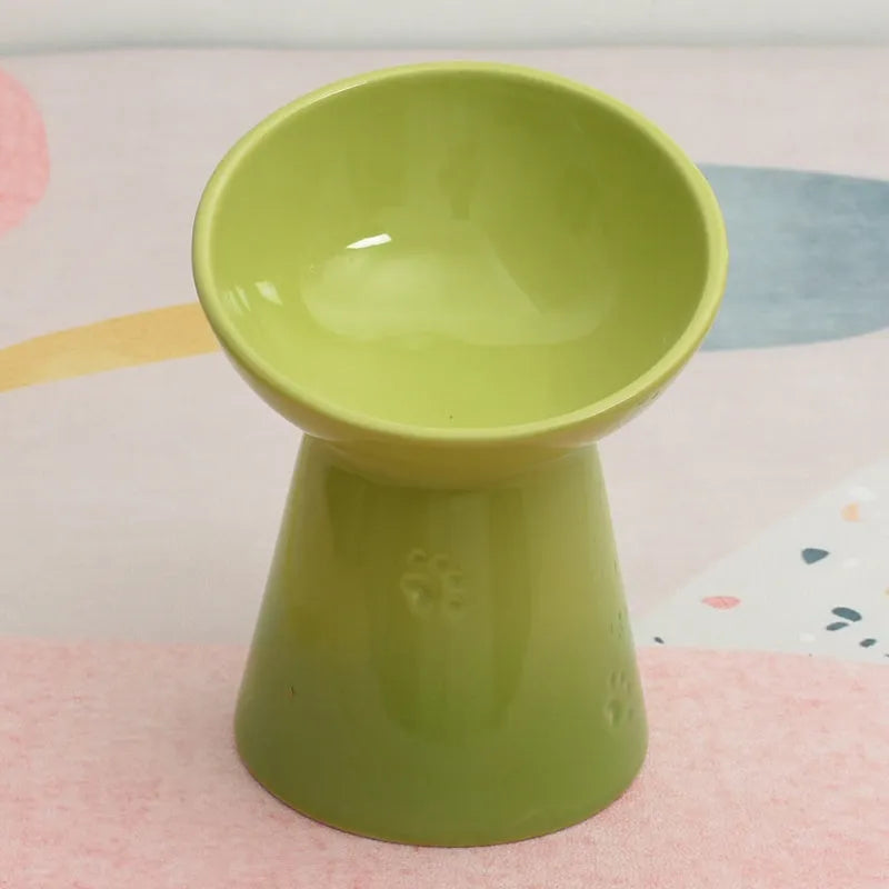 A high-footed white ceramic pet bowl with a slanted rim and non-slip black base, shown on a wooden floor.